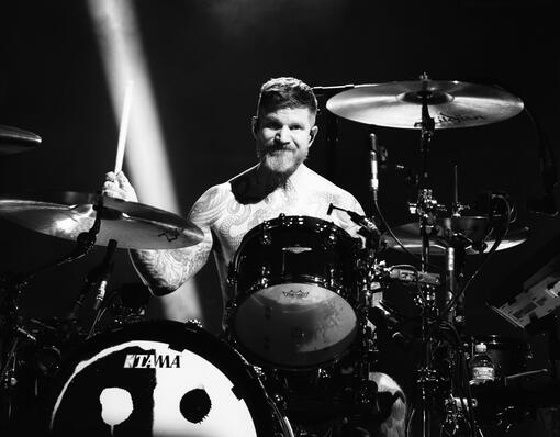 This is a photo of Andy Hurley playing drums, with the black and white half sad face, half smiley face new FOB logo on the bass drum.